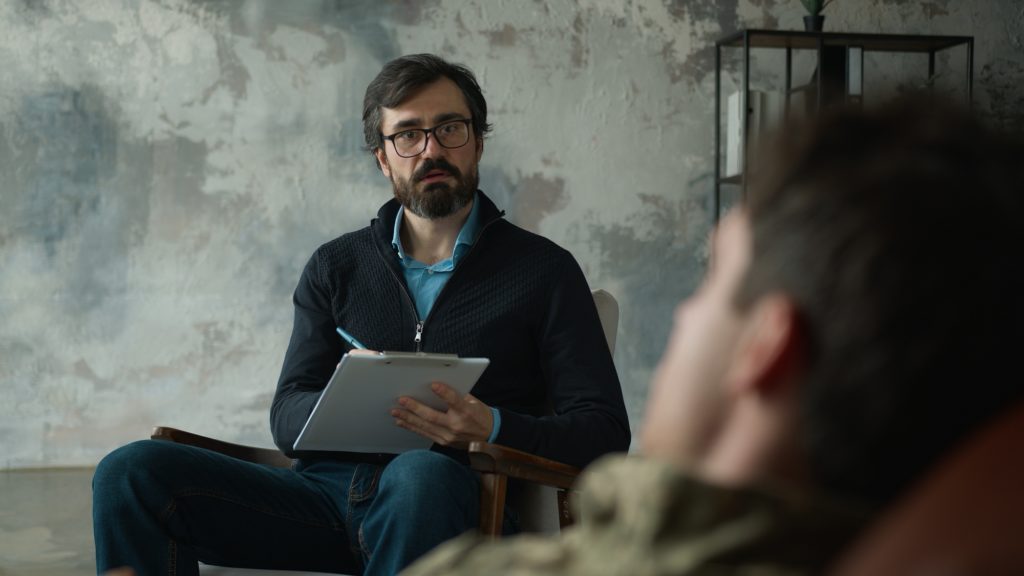 Worried mature Caucasian psychologist counselor writing notes listening patient client male officer